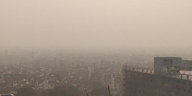 A hazy, smog-filled view of the cityscape in Delhi, with dense air pollution obscuring the buildings in the distance, creating a fog-like atmosphere